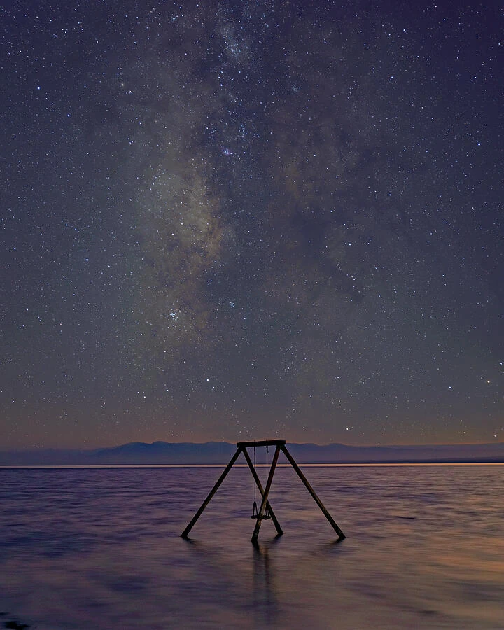 A partially submerged swing set beneath the Milky Way beside still water.