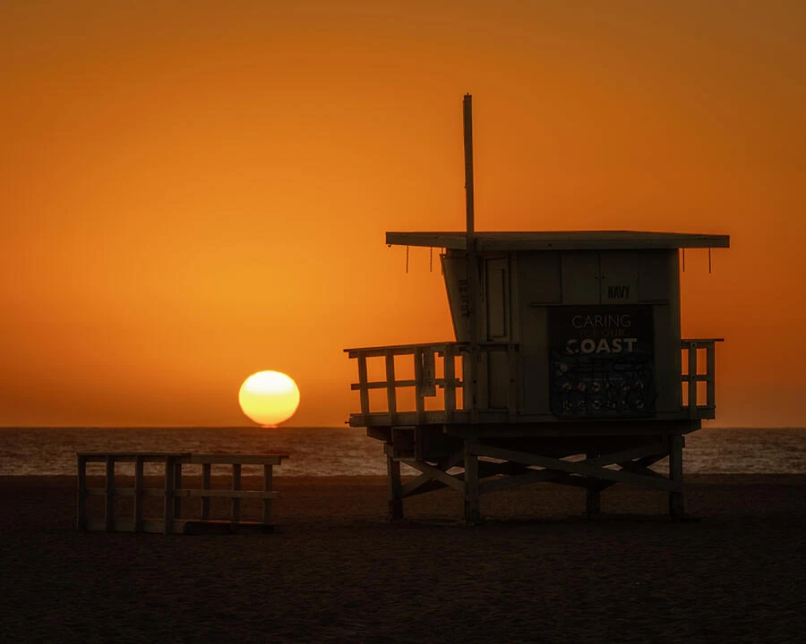 A lifeguard tower on the beach beneath a vivid orange sunset.